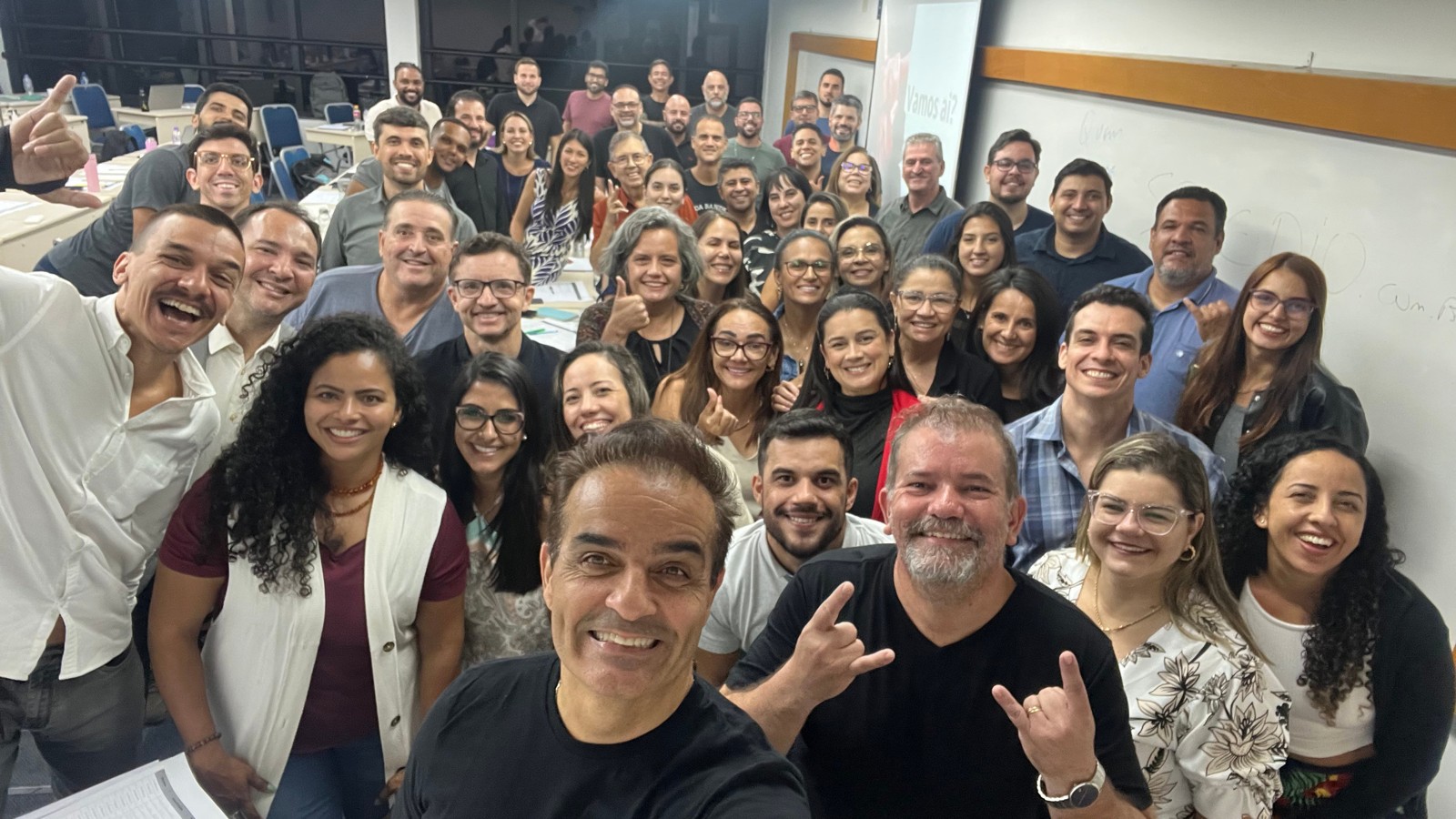 Jordão e uma turma reunida em uma sala de treinamento, fazendo gestos e sorrindo para a foto
